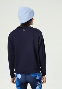 Marinblå sweatshirt med rund halsringning, ribbade ärmslut och en liten logopatch på ryggen. Matchas med blå leggings med blommönster och en ljusblå mössa.