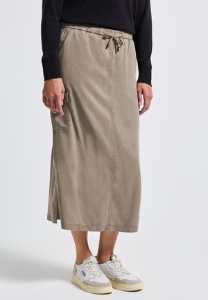 Nainen, jolla on beige midi-hame sivutaskuilla ja nyörivarsilla, musta pitkähiha on päällä ja valkoiset lenkkarit, seisoo valkoisella lattialla.