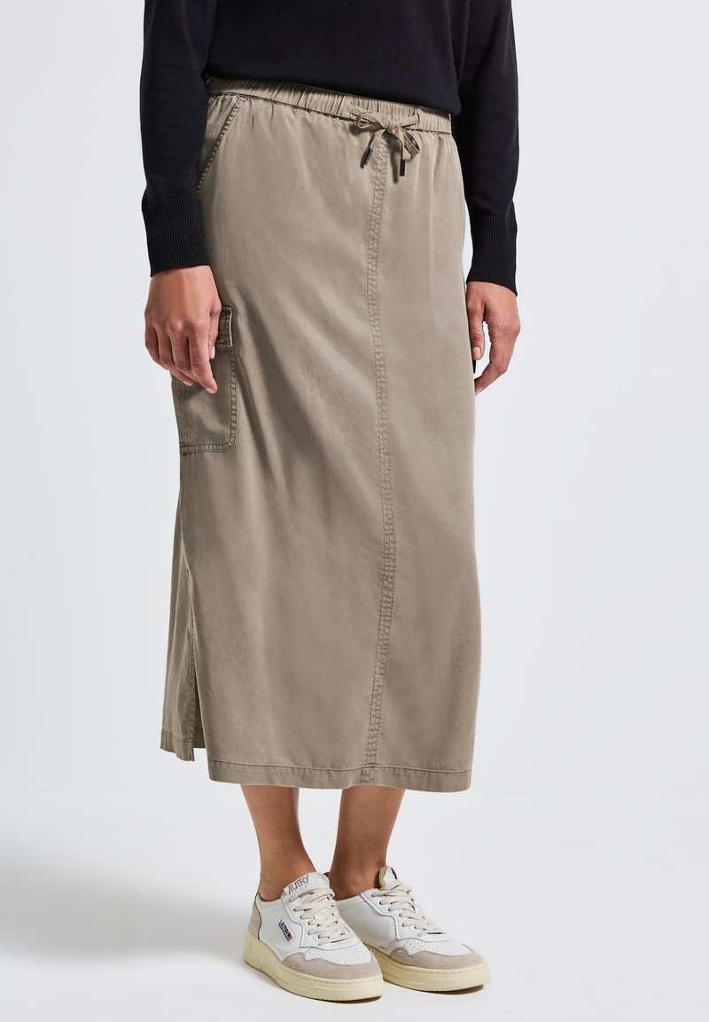 Nainen, jolla on beige midi-hame sivutaskuilla ja nyörivarsilla, musta pitkähiha on päällä ja valkoiset lenkkarit, seisoo valkoisella lattialla.