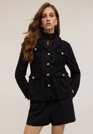 Zwarte op maat gemaakte blazer met gouden knopen, klepzakken en een gestructureerd ontwerp, gecombineerd met zwarte shorts en een kanten top met hoge hals.