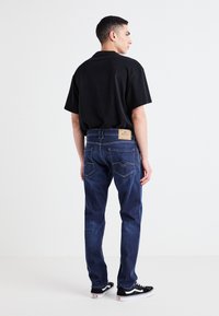 Mörkblå denimjeans med en klassisk passform, med en beige läderetikett bak, kombinerat med en svart bomullströja och svarta sneakers.