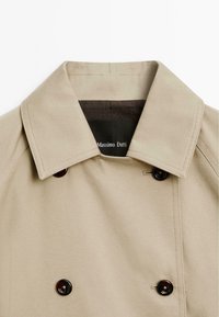 Cappotto trench beige doppiopetto con ampio colletto, caratterizzato da bottoni neri e una fodera marrone scuro, realizzato in tessuto liscio.