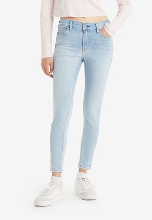 Person trägt hellblaue Skinny-Jeans mit hoher Taille und gekürztem Saum, kombiniert mit weißen Turnschuhen und Knöchelsocken vor einem einfarbigen Hintergrund.