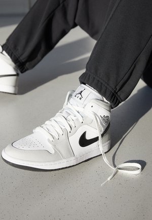Osoba w czarnych spodniach i butach Nike Air Jordan w kolorze białym i czarnym, jeden but rozwiązany i opierający się o jasnoszarą podłogę.