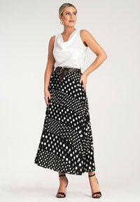 Biała satynowa bluzka bez rękawów, czarna plisowana spódnica w duże białe kropki oraz brązowy pasek. Modelka ma na sobie czarne sandały na obcasie.