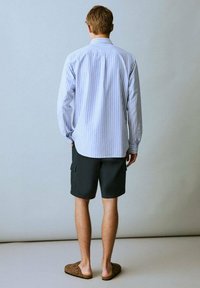 Un giovane uomo è in piedi con la schiena rivolta verso la camera, indossando una maglietta a righe con maniche lunghe, pantaloni corti scuri e pantofole casual su uno sfondo semplice.
