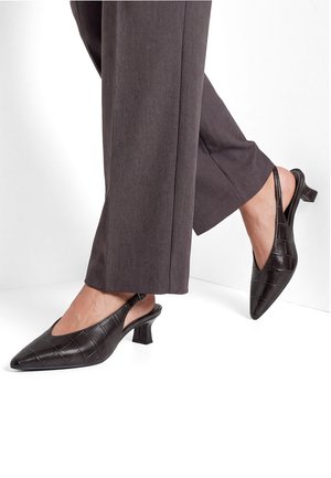 Donna che indossa pantaloni a gamba larga marrone scuro e scarpe slingback nere con motivo coccodrillo e tacco basso su sfondo bianco.
