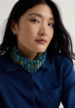 Giovane donna con lunghi capelli neri che indossa una camicia blu e una sciarpa al collo blu-verde a motivo, rivolta leggermente di lato contro uno sfondo semplice.