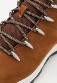 Botas de senderismo de ante marrón que presentan tela de malla marrón oscuro, ojales metálicos y cordones texturizados en verde y naranja, con una suela de goma blanca.