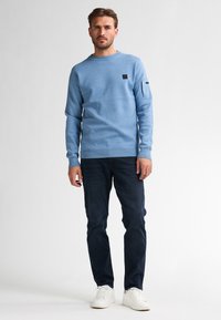 Lichtblauwe trui met ronde hals en zwart logoplaatje, gecombineerd met donkerblauwe jeans en witte sneakers. Zachte textuur en casual ontwerp.