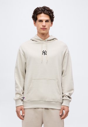 Fiatal férfi göndör sötét hajjal, bézs színű New York Yankees kapucnis pulóvert és hozzá illő melegítőnadrágot visel, egyszínű háttér előtt áll.
