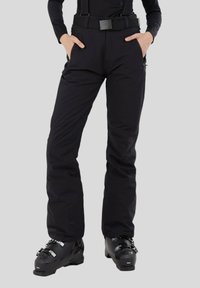 Schwarze Skihosen mit verstellbaren Trägern, Reißverschlusstaschen und strapazierfähigem Stoff; figurbetontes Design mit glatter Textur. Kombiniert mit schwarzen Skischuhen.