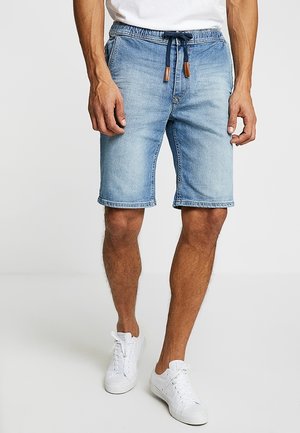 Mężczyzna w jasnoniebieskich szortach dżinsowych z troczkami i białych trampkach, stojący na jednolitym jasnym tle.