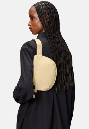 Donna con i capelli lunghi e intrecciati, indossa una giacca nera e porta una borsa a tracolla beige sopra la spalla su uno sfondo semplice.