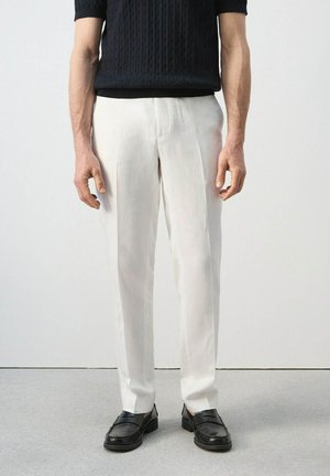 Hombre con pantalones blancos a medida, mocasines negros y una camisa negra de manga corta con textura, de pie sobre un suelo gris.