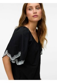 Blusa nera con maniche corte a sbuffo. Presenta dettagli in pizzo ricamato bianco lungo i bordi delle maniche. Tessuto dalla texture liscia, design informale.