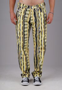 Man wearing yellow, black, and white striped pants with abstract patterns and black sneakers with white laces, standing against a gray background.