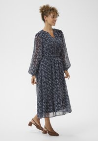 Kvinna med lockigt hår iförd en långärmad, marinblå blommig midiklänning och bruna klackskor med blockklack, stående och tittande åt sin högra sida.