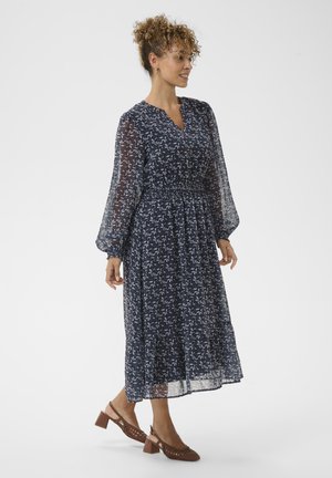 Ženska s kodrastimi lasmi, oblečena v dolgi rokav, mornarsko modro cvetlično midi obleko in rjavimi blok petkami, stoji in gleda v desno.