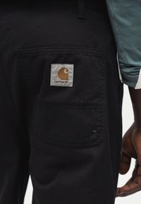 Κοντινό πλάνο τσέπης μαύρου παντελονιού Carhartt σε άτομο που φοράει τιρκουάζ πουκάμισο με μακριά μανίκια, εμφανίζοντας το διακριτικό λογότυπο Carhartt και μέρος του χεριού τους.