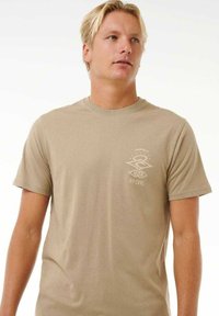 T-shirt beige a maniche corte in cotone con colletto rotondo, caratterizzato da un piccolo logo bianco di "RIP CURL" nella zona del petto.