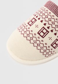 Papuc de culoare crem, realizat dintr-un material texturat, cu modele geometrice burgundy și un guler tricotat cu dungi. Formă moale, rotunjită.