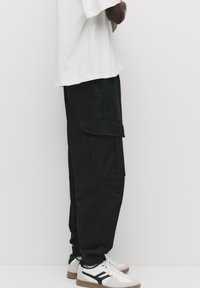 Zwarte cargo pants met een relaxte pasvorm, voorzien van meerdere zakken en elastische boorden. G gedragen met witte sneakers en een losse witte t-shirt.