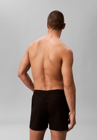 Kale man met kort haar, gezicht van ons af, draagt zwarte shorts met elastische taille en een achterzak, staand tegen een effen grijze achtergrond.