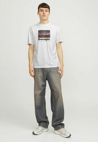 Weißes Baumwoll-T-Shirt mit einem grafischen Druck, der Palmen und das Wort "Overnight" zeigt. Kombiniert mit locker sitzenden, ausgewaschenen Denim-Jeans.
