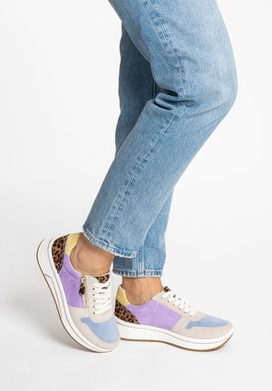 Persona che indossa jeans azzurri chiari e scarpe da ginnastica colorate con dettagli viola, blu, beige, giallo e stampa leopardata, in piedi su sfondo bianco.