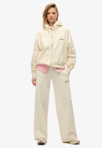 Sweat à capuche couleur crème avec fermeture éclair à l'avant, cordon de serrage ajustable et logo brodé. Associé à un pantalon large assorti. Tissu doux et texturé.