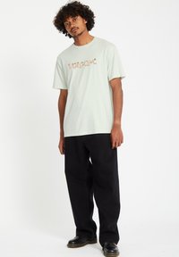 Ljusgrön bomullst-shirt med en stiliserad "Volcom"-logotyp i kontrasterande färger, tillsammans med svarta vida jeans och svarta skor.