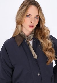 Ung kvinde med langt bølget blondt hår iført en navy jakke med brun krave og et beige mønstret halstørklæde.