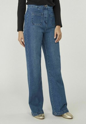Vrouw draagt een blauwe high-waist wijde jeans met voorvakken, gecombineerd met een zwarte top met lange mouwen en gouden schoenen.