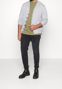 Svetle šedý cardigan s tmavými knoflíky, olivově zelené tričko pod tím, černé slim-fit džíny a černé chelsea boty. Jednoduchý design, volný střih.