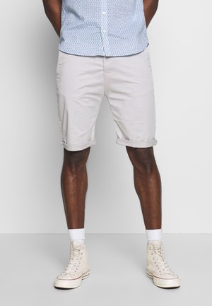 Jasnokremowe szorty wykonane z gładkiego materiału, z podwiniętymi mankietami. W połączeniu z białymi skarpetkami i beżowymi sneakersami za kostkę.