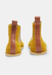 Botas Chelsea de ante amarillo con acentos en cuero marrón y tiradores, que presentan una superficie texturizada y detalles de costura.