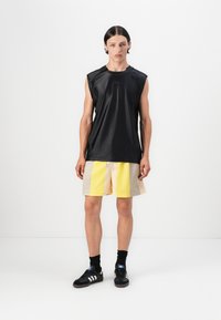 Svart ermeløs topp med glatt tekstur, paret med gule og beige mesh shorts. Modellen har på svarte joggesko med hvite striper og sokker.
