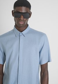 Camicetta azzurra chiara a maniche corte, con colletto, superficie liscia e vestibilità slim. Occhiali da sole neri angolari coprono gli occhi.