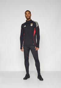 Zwarte sportieve trainingsoutfit met rode accenten, rits bovenkleding en lange mouwen, gecombineerd met strakke broeken. Bevat het Benfica-logo en drie strepen.