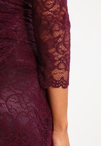 Gros plan sur une manche de robe en dentelle bordeaux montrant des motifs floraux et un bord festonné avec la peau visible en dessous.