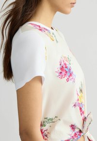 Blomsterprintet top med hvide korte ærmer, rund halsudskæring og en knude detalje i taljen, med rosa, gul og grønne accenter.
