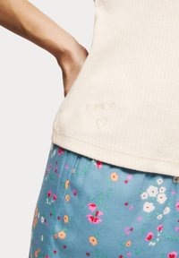 Krämfärgad ribbad linne med broderad "love" och hjärtärmne vid nederkanten, bärs tillsammans med blå blommönstrade byxor med olika färgade blommor.