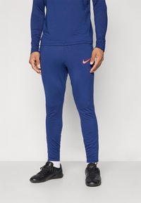 Jūras zila sporta bikses ar pieguļošu dizainu, ar oranžu Nike logo uz labā kāja. Uzvilktas kopā ar melniem sporta apaviem.