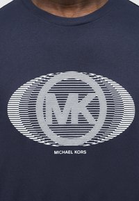 Μπλε πουκάμισο με λευκό οβάλ ριγέ μοτίβο που περιλαμβάνει μεγάλα αρχικά "MK" και κείμενο "Michael Kors" κάτω από το σχέδιο.