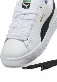 Baskets blanches Puma Suede XL avec bande noire, lacets épais blancs, logo doré sur le côté, et lacets noirs supplémentaires à côté de la chaussure.