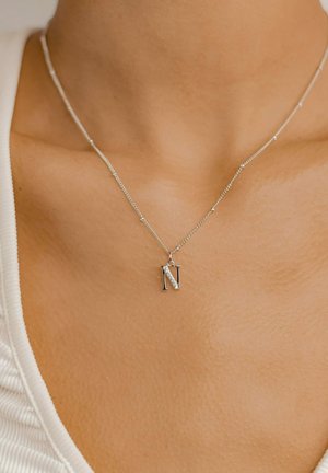 Silver necklace featuring an 'N' pendant with a diagonal line of clear stones, on a delicate chain with small beads. Skin tone visible.