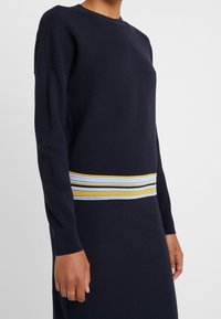 Maglione lavorato a maglia blu navy con tessuto strutturato, caratterizzato da una fascia a righe gialla, bianca e blu in vita. Girocollo, maniche lunghe.
