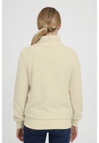 Mujer con cabello rubio en una coleta, vistiendo una chaqueta esponjosa color crema y jeans azules, vista desde atrás contra un fondo lizo.