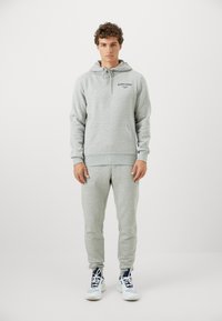 Mężczyzna stoi w jasnoszarej bluzie z kapturem i dopasowanych dresach, w sportowych butach, na tle jednolitego białego tła.